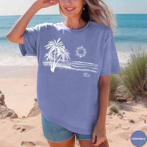 May include: A periwinkle blue short-sleeved t-shirt featuring a white line-art graphic of three palm trees, a sun, ocean waves, and a small seashell. The shirt has a crew neckline and a relaxed fit, showcasing a coastal-themed illustration.