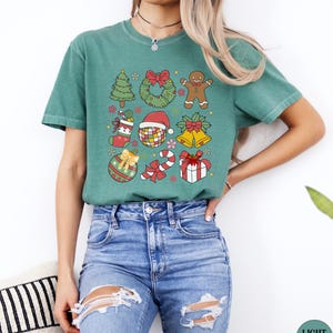 May include: A sage green short-sleeve t-shirt featuring a graphic print of various Christmas icons including a tree, wreath, gingerbread person, stocking, disco ball with a Santa hat, bells, ornament, candy cane, and a gift box with a red bow.