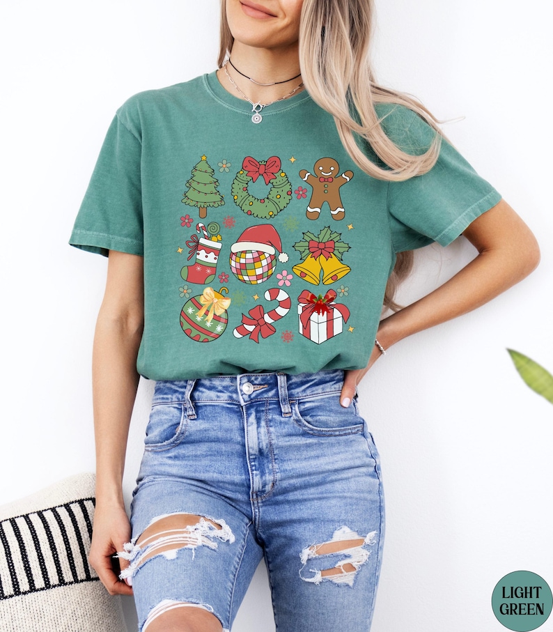 May include: A sage green short-sleeve t-shirt featuring a graphic print of various Christmas icons including a tree, wreath, gingerbread person, stocking, disco ball with a Santa hat, bells, ornament, candy cane, and a gift box with a red bow.