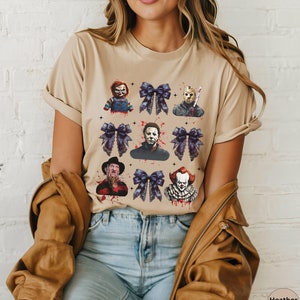 May include: Heather tan t-shirt featuring a graphic design of horror movie characters and dark purple bows. The design includes Chucky, Jason Voorhees, Michael Myers, Freddy Krueger, and Pennywise. The shirt is a neutral color.