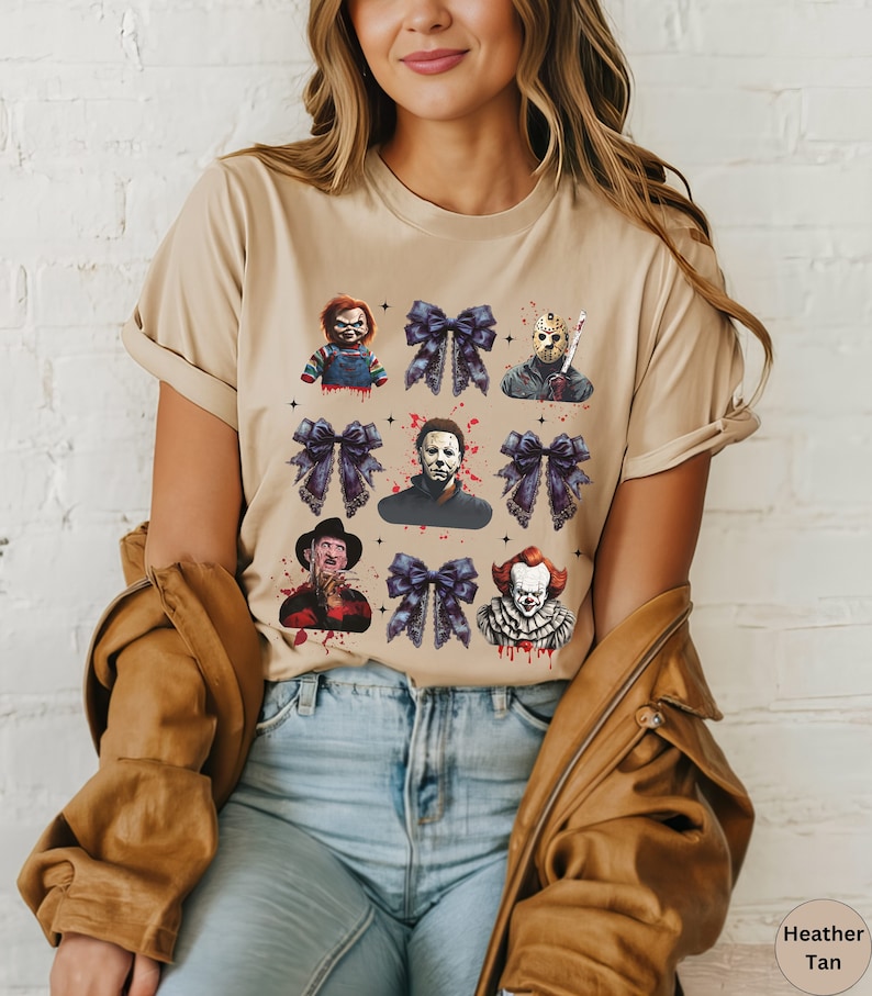 May include: Heather tan t-shirt featuring a graphic design of horror movie characters and dark purple bows. The design includes Chucky, Jason Voorhees, Michael Myers, Freddy Krueger, and Pennywise. The shirt is a neutral color.