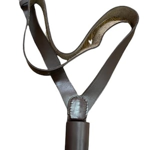 Puede incluir: Una correa de cámara de cuero marrón con una hebilla dorada. La correa tiene un diseño único con un lazo para el cuello y una pieza cilíndrica en la parte inferior. El cuero parece ser de alta calidad.