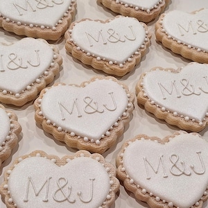 May include: Heart-shaped cookies with white icing and the letters "M&J" in a delicate script. The cookies have a scalloped edge and are decorated with small white pearls around the border. A sweet treat.