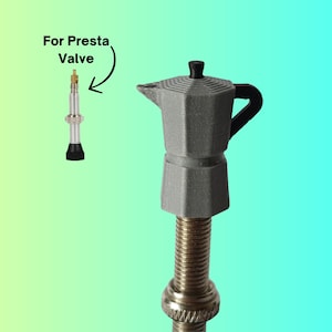 Op de afbeelding: Een grijze, 3D-geprinte moka-pot ventieldop bevestigd aan een metalen Presta-ventiel. De dop heeft een zeshoekig lichaam, een zwarte handgreep en een zwarte knop op het deksel. De tekst "For Presta Valve" staat met een pijl bij het ventiel.