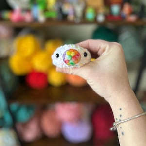 May include: A white crocheted eye-shaped plush toy with a colourful, round centre. The toy has black button eyes and is held up against a blurred background of other colourful crocheted items. The plush is handmade.