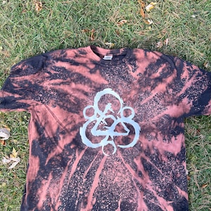 Può includere: Una t-shirt tie-dye nera e marrone con un design sbiancato bianco di un cerchio con un triangolo all'interno. Il design ricorda il logo di una band.