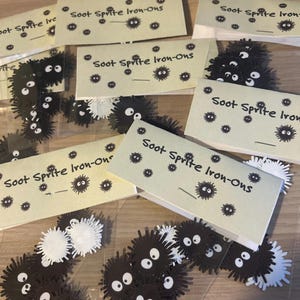 May include: A clear plastic bag filled with black and white iron-on patches featuring a cartoon character with large eyes. The patches are labelled "Soot Sprite Iron-Ons".