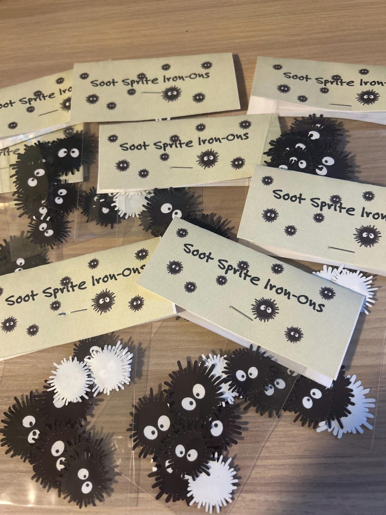 May include: A clear plastic bag filled with black and white iron-on patches featuring a cartoon character with large eyes. The patches are labeled "Soot Sprite Iron-Ons".