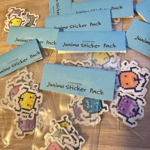 Puede incluir: Un conjunto de paquetes de pegatinas Junimo, cada uno con personajes pixelados coloridos. Las pegatinas están empaquetadas en bolsas de plástico transparentes con etiquetas azules que dicen "Junimo Sticker Pack".
