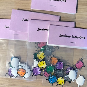 May include: A clear plastic bag filled with colorful iron-on patches featuring pixelated characters. The patches are shaped like small, cartoon-like creatures with different colors and designs. The bag is labeled "Junimo Iron-Ons".