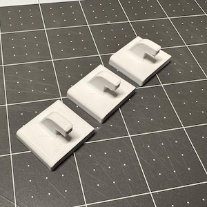 May include: Three light gray, square-shaped 3D-printed objects with a curved, rectangular handle extending from one side. The objects are arranged diagonally on a dark gray surface with a grid pattern.