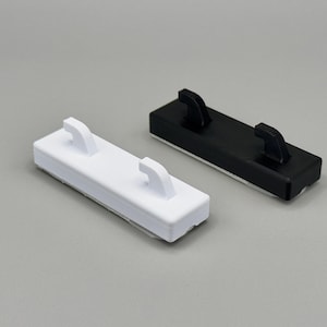 Peut inclure: Deux crochets muraux rectangulaires, l'un blanc et l'autre noir. Chaque crochet pr&eacute;sente une base plate avec deux tiges incurv&eacute;es orient&eacute;es vers le haut, con&ccedil;ues pour maintenir des objets en toute s&eacute;curit&eacute; contre une surface plane.