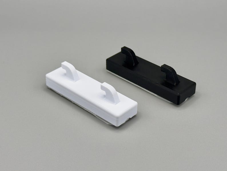 Peut inclure: Deux crochets muraux rectangulaires, l'un blanc et l'autre noir. Chaque crochet pr&eacute;sente une base plate avec deux tiges incurv&eacute;es orient&eacute;es vers le haut, con&ccedil;ues pour maintenir des objets en toute s&eacute;curit&eacute; contre une surface plane.