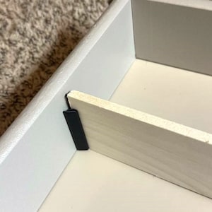 May include: Close-up of a partially assembled white drawer. The drawer's interior is light beige, with a wooden divider and a black corner bracket. The drawer is set against a neutral-colored carpet.