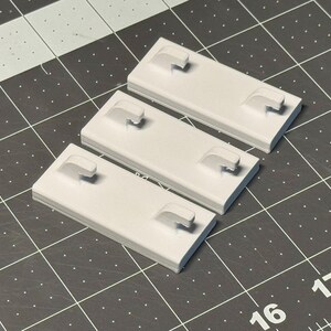 May include: Three light gray rectangular plastic components with a curved handle-like protrusion on each. The objects are arranged on a dark gray cutting mat with a grid pattern and white markings.