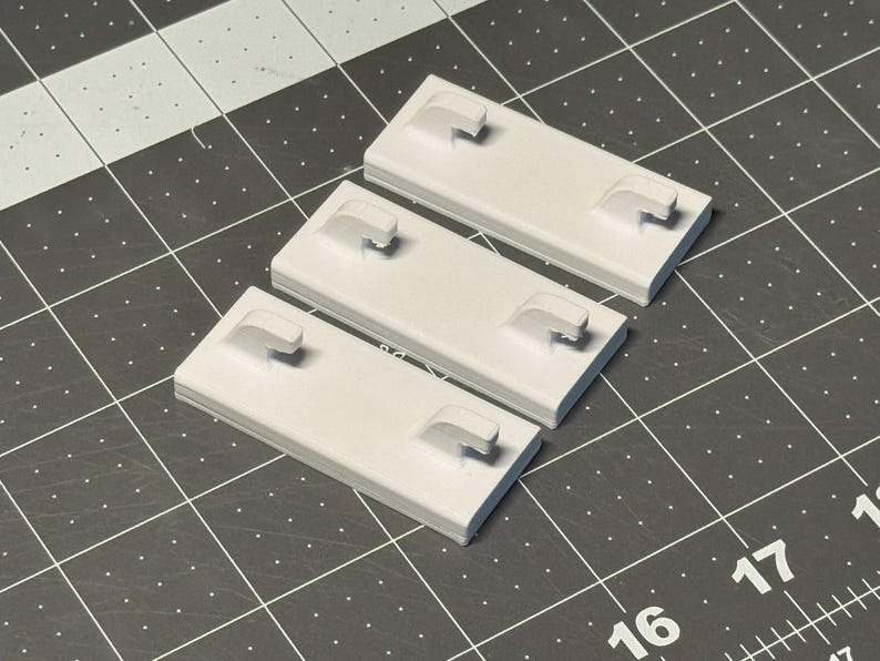 May include: Three light gray rectangular plastic components with a curved handle-like protrusion on each. The objects are arranged on a dark gray cutting mat with a grid pattern and white markings.