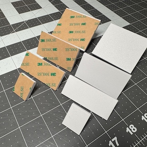 May include: An assortment of white foam pads and brown adhesive squares with the text "3M 300LSE" and "OLSE" printed on them. The items are arranged on a black cutting mat with white grid lines.