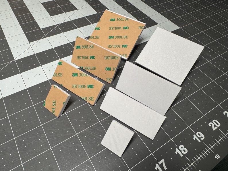 May include: An assortment of white foam pads and brown adhesive squares with the text "3M 300LSE" and "OLSE" printed on them. The items are arranged on a black cutting mat with white grid lines.