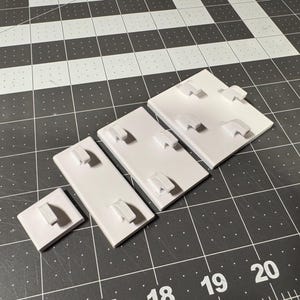 May include: A collection of white jewelry display cards in various sizes, each featuring a raised, curved holder. The cards are arranged on a black and white grid surface, providing a clean and organized presentation for showcasing earrings or other small jewelry items.