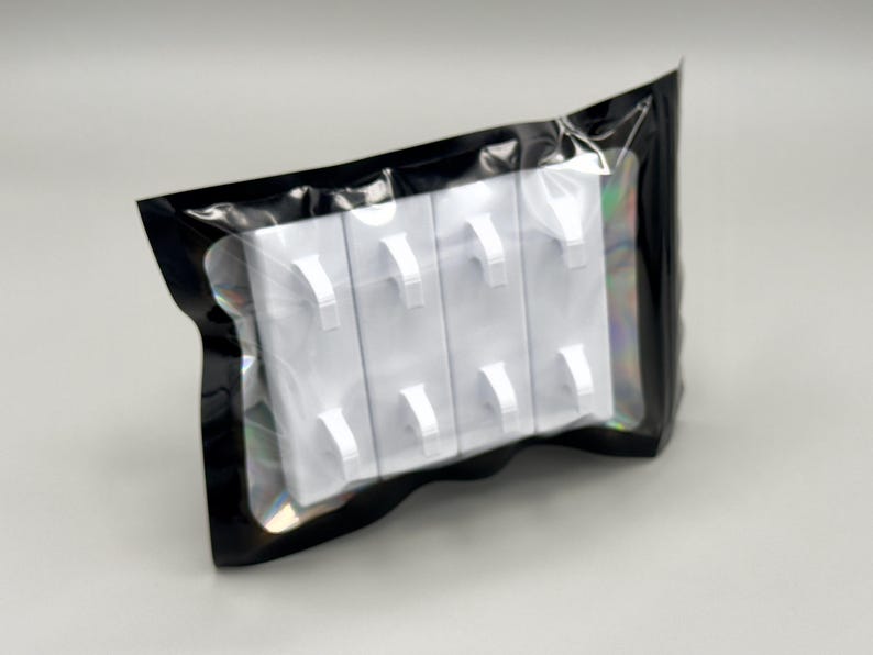Peut inclure: Un ensemble de quatre crochets muraux rectangulaires en plastique blanc, chacun dot&eacute; de deux tiges incurv&eacute;es, scell&eacute; dans une pochette en plastique noir avec une fen&ecirc;tre transparente qui r&eacute;v&egrave;le le contenu &agrave; travers la face avant de l'emballage.