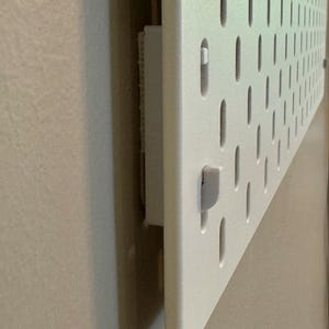 May include: A white pegboard with evenly spaced, oval-shaped holes. A white, rectangular object is attached to the back of the board. A small, white peg is inserted into one of the holes. The pegboard is mounted on a light-colored wall.