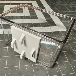 May include: A clear, rectangular plastic container with a gray, 3D-printed bracket attached. The bracket has four curved hooks. The container sits on a black and white checkered surface.
