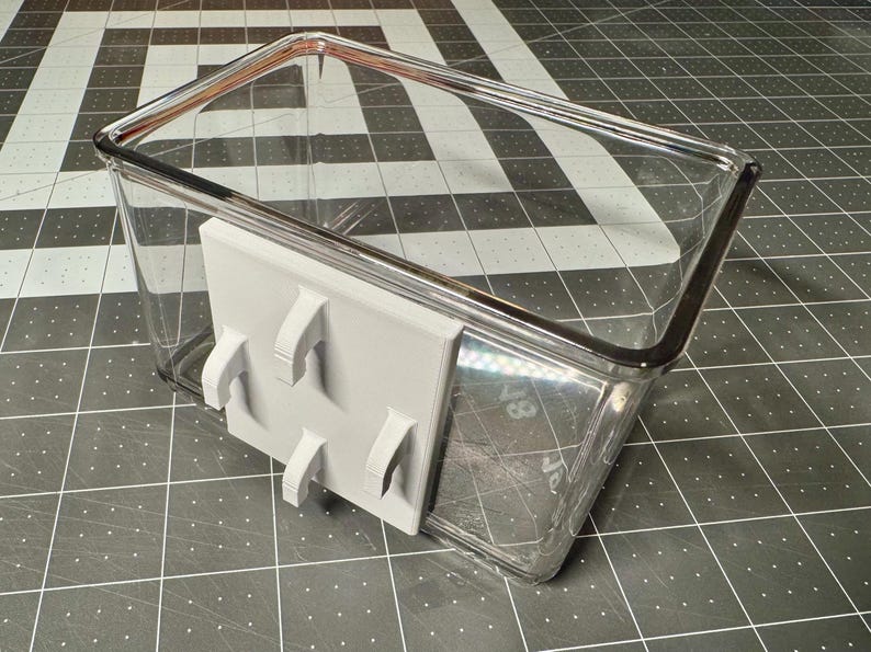 May include: A clear, rectangular plastic container with a gray, 3D-printed bracket attached. The bracket has four curved hooks. The container sits on a black and white checkered surface.