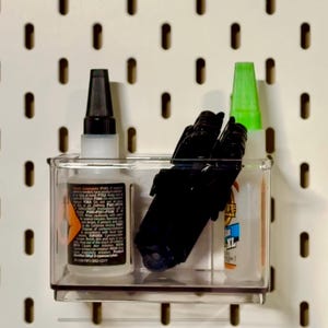 May include: A clear plastic container holding various items, including a bottle of glue with a black cap, several black markers, and a bottle with a green cap. The container is mounted on a white pegboard with evenly spaced holes.