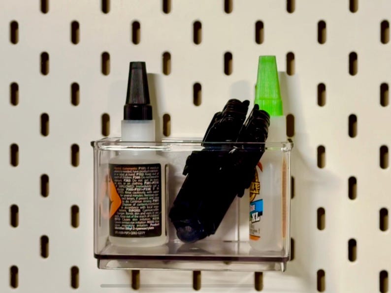 May include: A clear plastic container holding various items, including a bottle of glue with a black cap, several black markers, and a bottle with a green cap. The container is mounted on a white pegboard with evenly spaced holes.