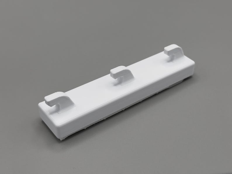 Peut inclure: Une bande de crochet murale rectangulaire en plastique blanc dot&eacute;e de trois crochets uniform&eacute;ment espac&eacute;s et incurv&eacute;s vers le haut. La base est un bloc solide et allong&eacute; avec une finition lisse, con&ccedil;ue pour &ecirc;tre fix&eacute;e sur une surface plane.