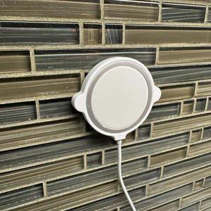 May include: A white, circular wireless charger with a silver trim and a white cord. The charger is mounted on a mosaic tile wall with brown, gray, and beige tiles. The charger has a minimalist design.