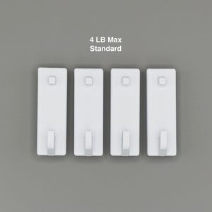 Peut inclure: Quatre crochets adh&eacute;sifs rectangulaires blancs dispos&eacute;s en ligne. Chaque crochet pr&eacute;sente un petit bouton carr&eacute; pr&egrave;s du sommet et un crochet courb&eacute; &agrave; la base. Le texte 4 LB Max Standard est inscrit au-dessus, indiquant la capacit&eacute; de poids.