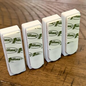 Peut inclure: Quatre crochets muraux adh&eacute;sifs rectangulaires en plastique blanc dispos&eacute;s en rang&eacute;e. Chaque crochet pr&eacute;sente une bande adh&eacute;sive en mousse blanche au dos avec le logo vert de la marque Command imprim&eacute; &agrave; plusieurs reprises sur la surface.