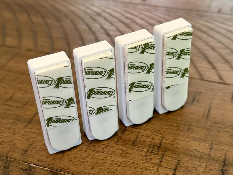 Peut inclure: Quatre crochets muraux adh&eacute;sifs rectangulaires en plastique blanc dispos&eacute;s en rang&eacute;e. Chaque crochet pr&eacute;sente une bande adh&eacute;sive en mousse blanche au dos avec le logo vert de la marque Command imprim&eacute; &agrave; plusieurs reprises sur la surface.