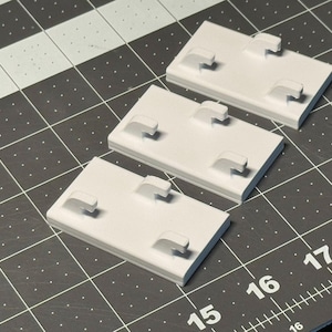 May include: Three white rectangular plastic components with four raised hooks each. The components are arranged on a black and gray grid surface, with measurements visible along the edges. The hooks are evenly spaced on each component.