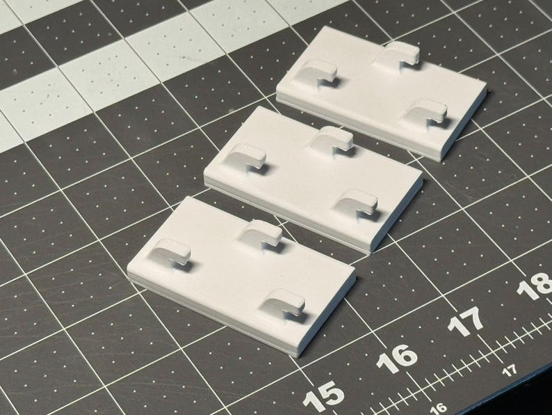 May include: Three white rectangular plastic components with four raised hooks each. The components are arranged on a black and gray grid surface, with measurements visible along the edges. The hooks are evenly spaced on each component.