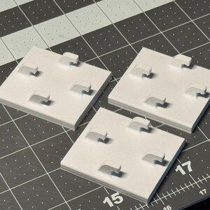 May include: Three white square jewelry display stands with four curved display arms each. The stands are arranged on a dark gray cutting mat with a grid pattern. The stands are made of a solid material.