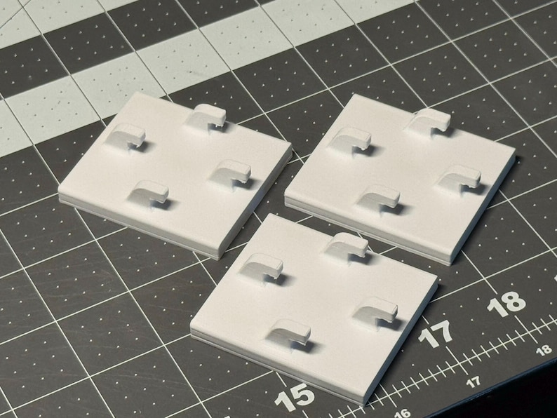May include: Three white square jewelry display stands with four curved display arms each. The stands are arranged on a dark gray cutting mat with a grid pattern. The stands are made of a solid material.