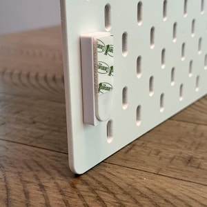 Peut inclure: Un panneau perfor&eacute; rectangulaire blanc dot&eacute; d'une grille de fentes ovales verticales. Une bande de montage adh&eacute;sive blanche avec la marque verte Command est fix&eacute;e sur le bord lat&eacute;ral du panneau pour faciliter l'installation murale.