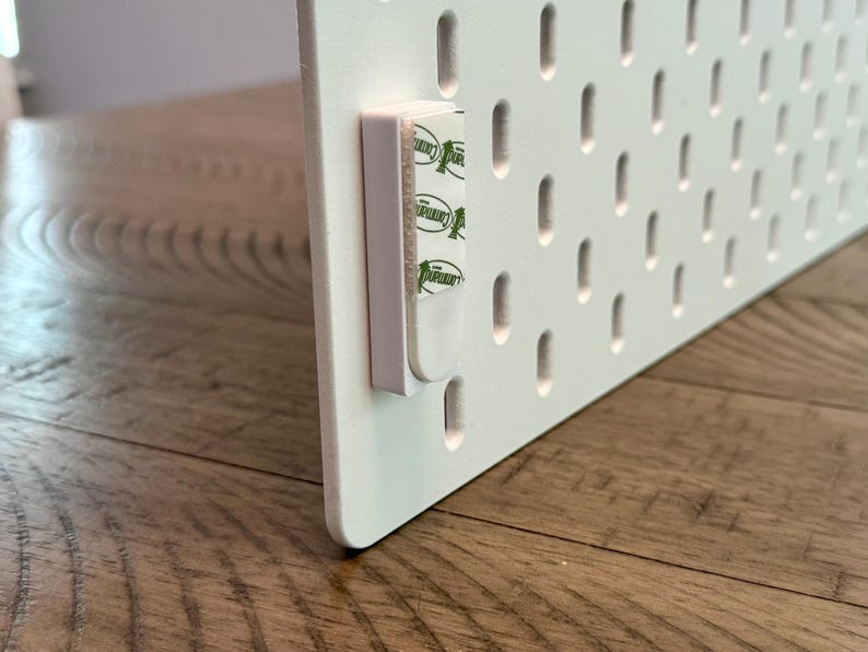 Peut inclure: Un panneau perfor&eacute; rectangulaire blanc dot&eacute; d'une grille de fentes ovales verticales. Une bande de montage adh&eacute;sive blanche avec la marque verte Command est fix&eacute;e sur le bord lat&eacute;ral du panneau pour faciliter l'installation murale.