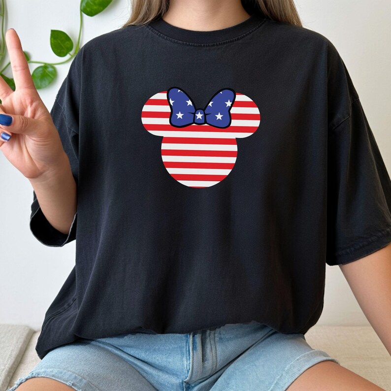 May include: Black t-shirt with a Minnie Mouse-style design. The design features red and white stripes and a blue bow with white stars. The design is centered on the front. The shirt has a relaxed fit.
