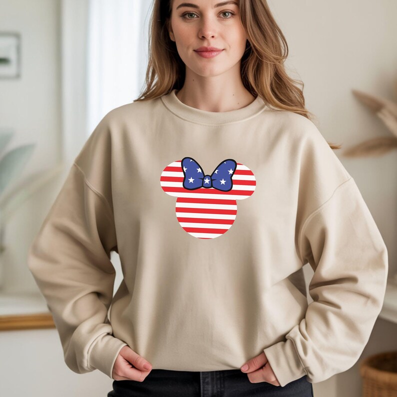 May include: Beige crew neck sweatshirt with a patriotic Minnie Mouse design. The design features a red and white striped head with a blue bow decorated with white stars. The sweatshirt is a neutral colour, suitable for casual wear.