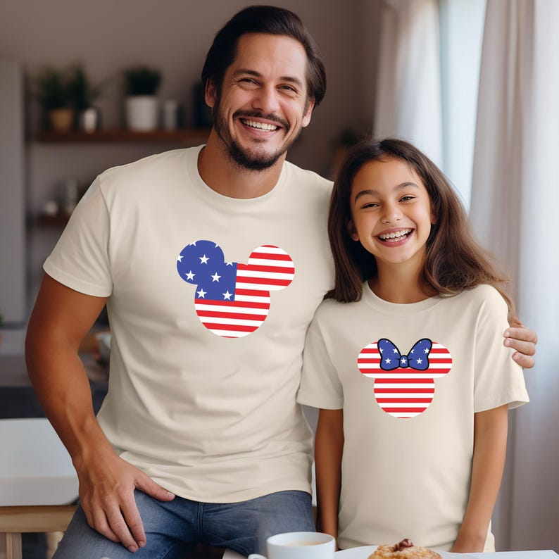 May include: Two matching beige t-shirts, one for an adult and one for a child. Each shirt features a Mickey Mouse head design with the American flag. The adult shirt has the full design, while the child's has a bow.