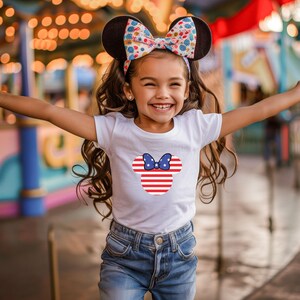 May include: A young girl wearing a white t-shirt featuring a Minnie Mouse design in red, white, and blue. She is also wearing a headband with Minnie Mouse ears and a colourful bow. The girl is smiling with her arms outstretched.