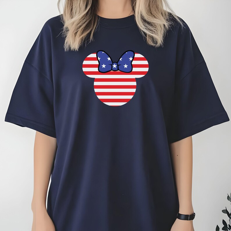 May include: Navy blue t-shirt with a Minnie Mouse design. The design features red and white stripes and a blue bow with white stars. The shirt has short sleeves and a crew neck. The design is centered on the front.