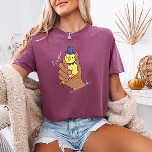 May include: A maroon t-shirt with a graphic of a yellow chick wearing a blue top hat and bow tie, held in a brown hand. The text "Sup chick" is written in white script. The model is wearing denim shorts.