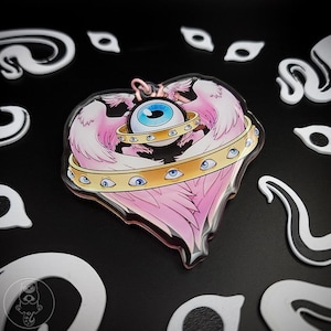 May include: A heart-shaped acrylic charm featuring a central blue eye surrounded by a gold band with smaller blue eyes. Pink feathered wings and a gold halo complete the design. White and black decorative shapes surround the charm.