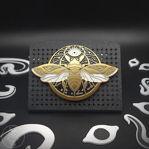 May include: A gold and white metal bee pin with an eye symbol on a black background. The bee has detailed wings and is set against a circular design. The pin is mounted on a black, perforated surface.
