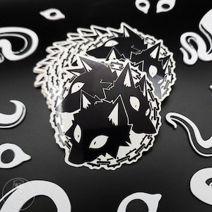 May include: A stack of black and white stickers featuring a wolf head design with a chain around the neck. The stickers have a glossy finish and are surrounded by other white decorative shapes on a black background.