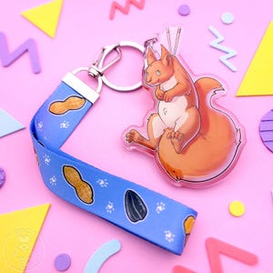 May include: A keychain with a cartoon squirrel charm and a blue lanyard. The squirrel is orange and white, holding chopsticks. The lanyard has a peanut and sunflower seed pattern. The keychain is on a pink background with colorful shapes.